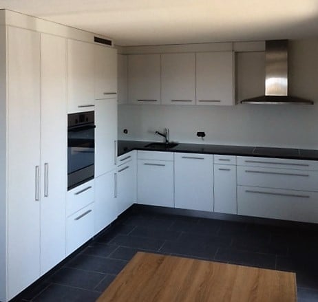 Modern kitchen with white cabinets, black countertops, and a dark tiled floor. Features include a sink, oven, and a range hood.
