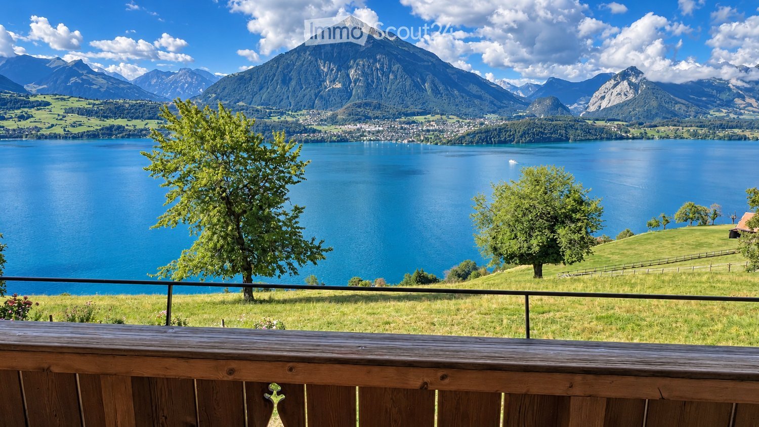 Balcony overlooking a serene lake surrounded by lush green hills and distant snow-capped mountains.