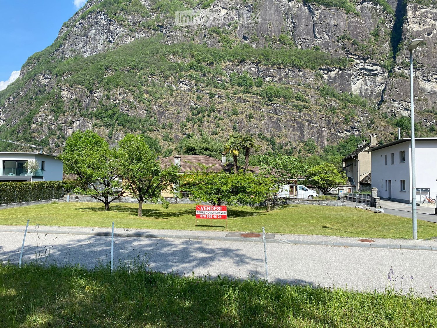 A well-maintained lawn with trees and a sign advertising a property for sale, with a large rocky mountain in the background.