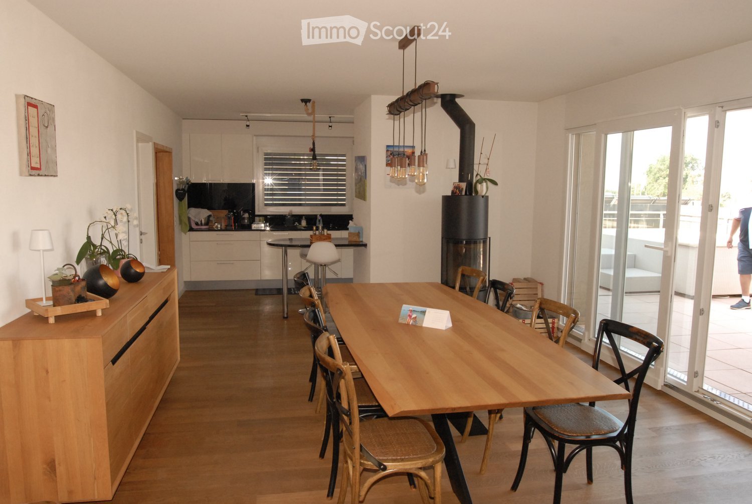 Open-plan living and dining area with a large wooden dining table, chairs, kitchen area with cabinets, a hanging light fixture, and a fireplace.