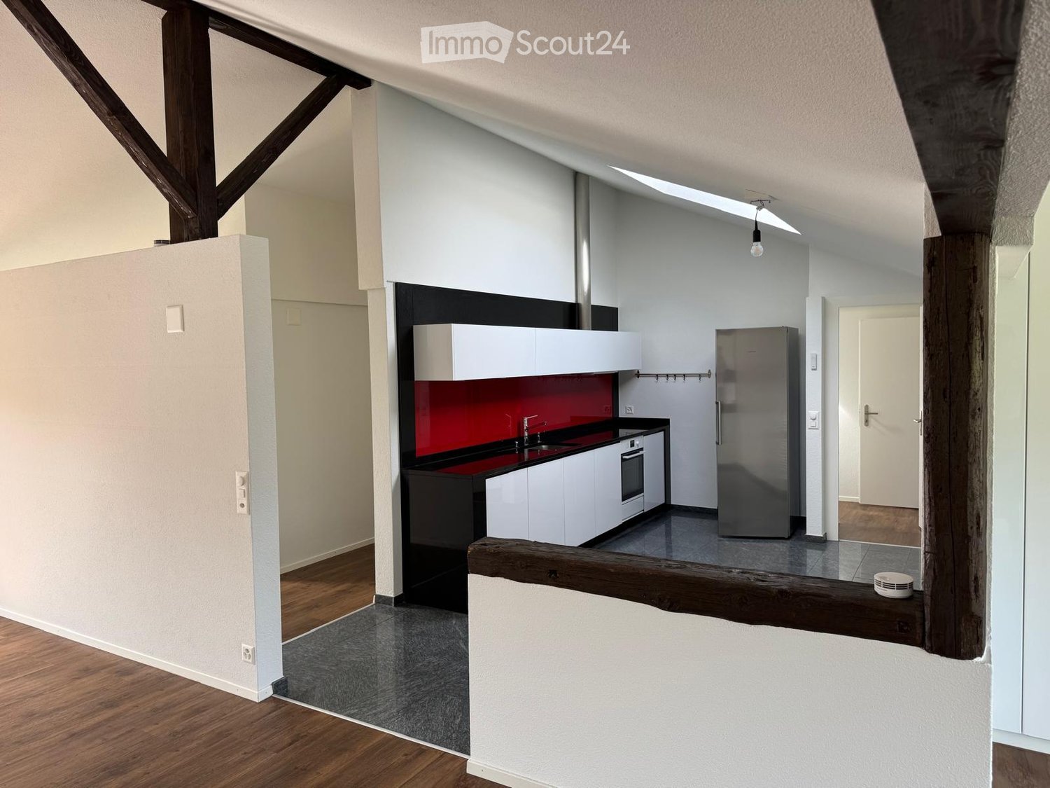 Modern kitchen with white cabinets, stainless steel appliances, and a red backsplash. The space features wooden flooring and exposed wooden beams.