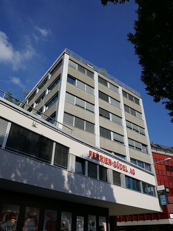 Multi-story building with a modern facade, large windows, and a sign reading "FERRIER-GUDEL-AG" on the lower level.
