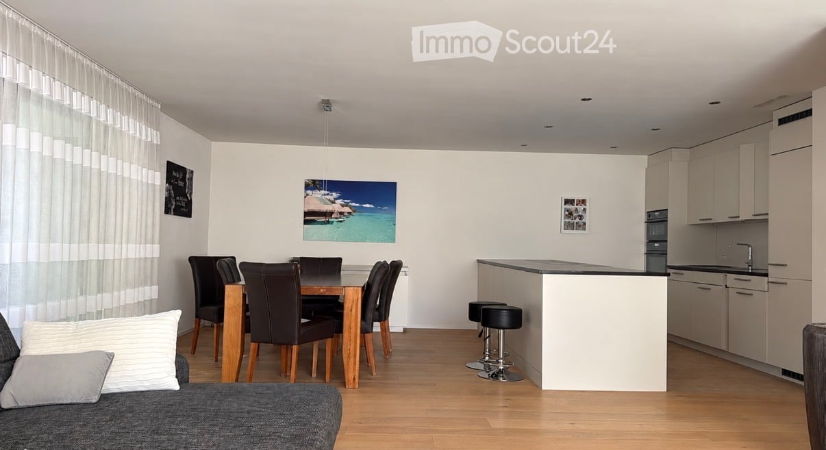 Open-plan living and dining area with a grey sofa, dining table with six chairs, kitchen island with stools, and modern cabinets.