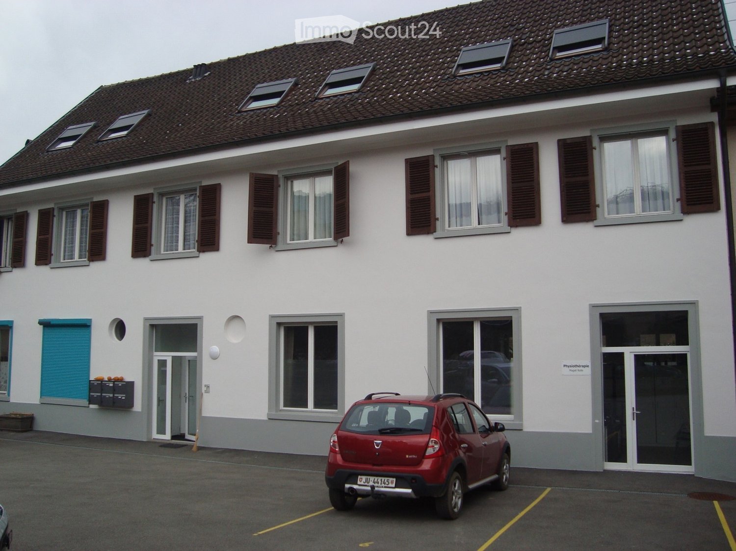 Two-story building with white exterior, multiple windows with shutters, and a red car parked in front.