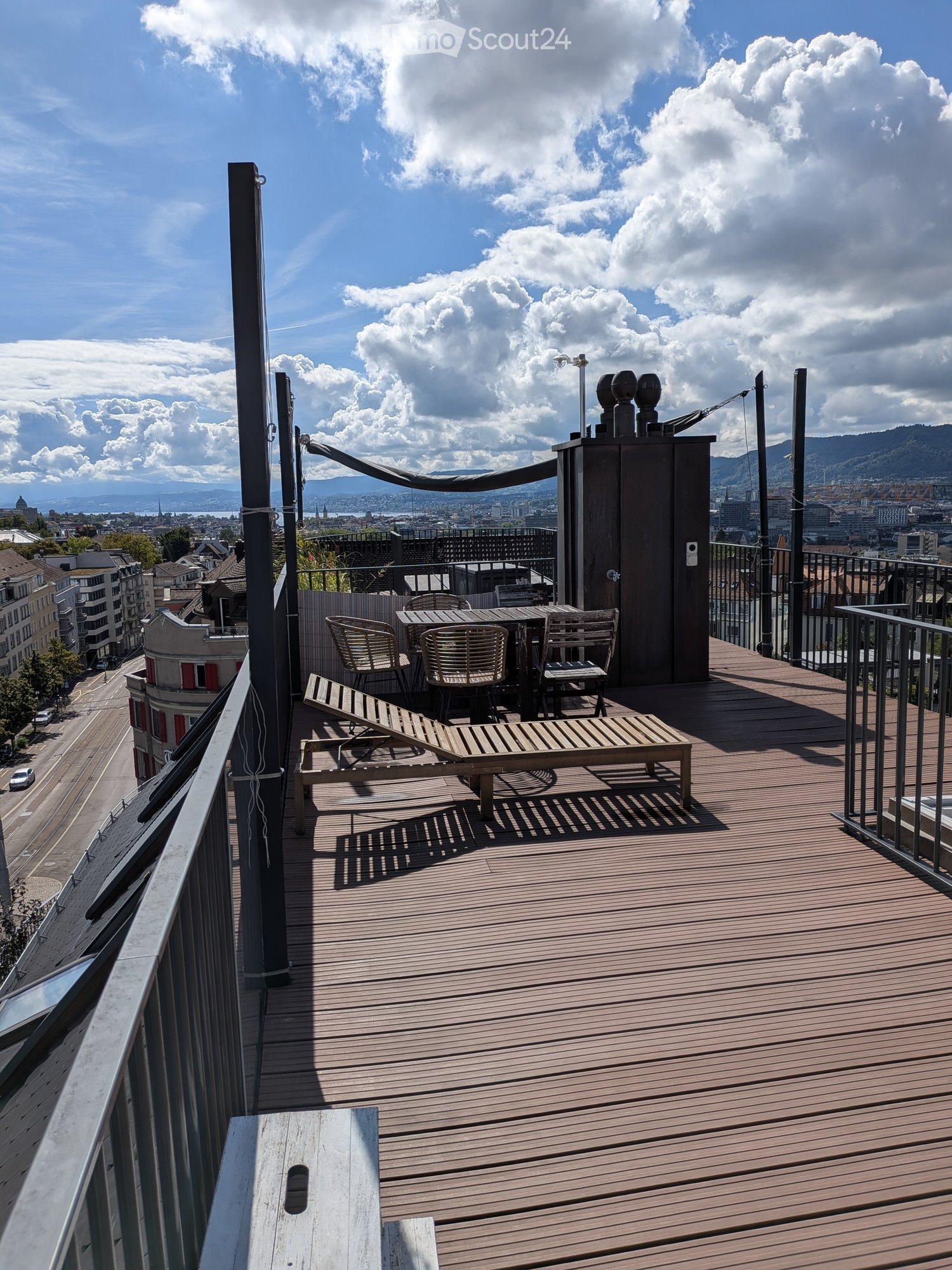 Wooden deck terrace with a table, chairs, and a lounge chair, overlooking a cityscape with mountains in the background.
