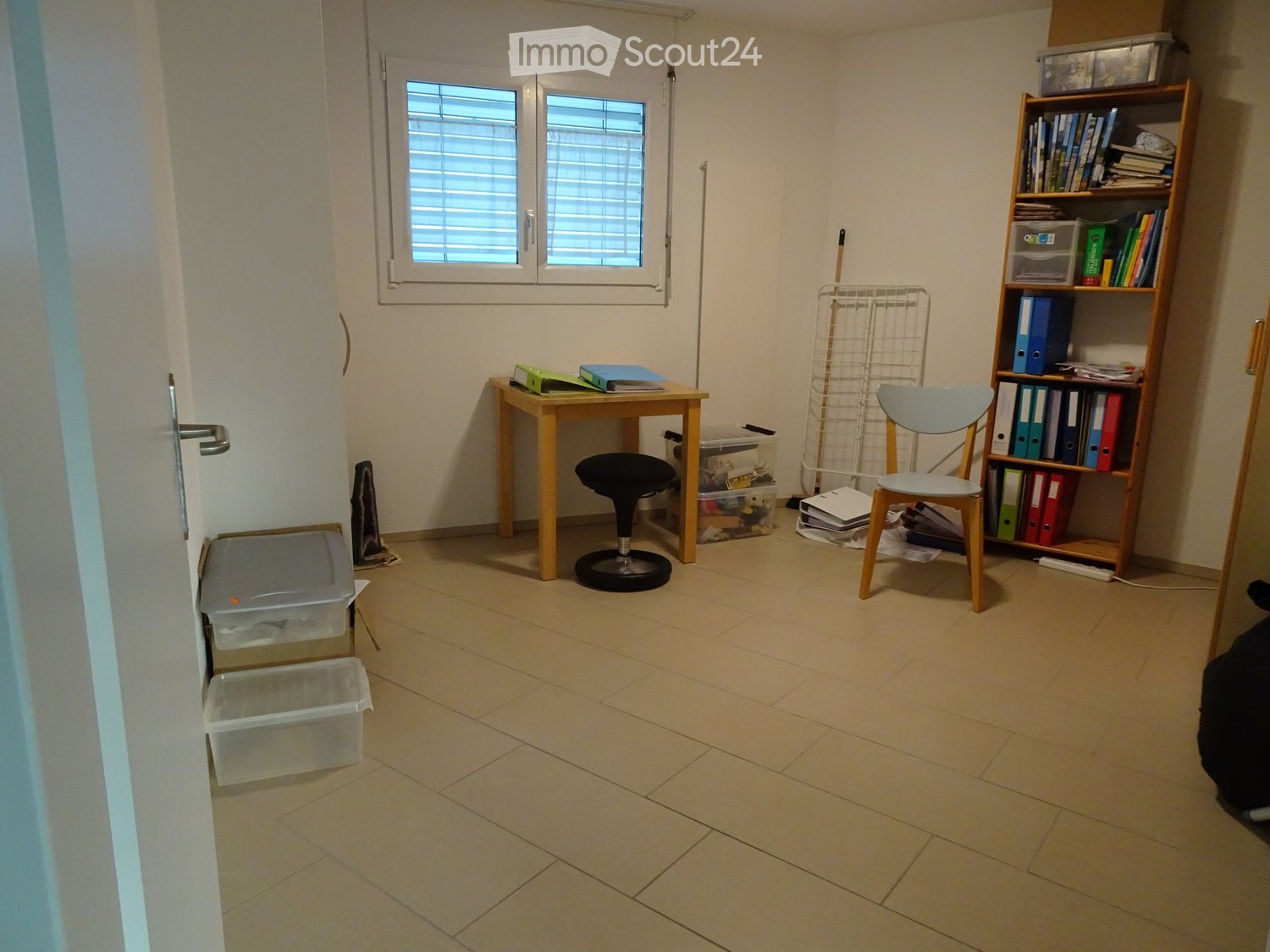 A small, furnished room with a tiled floor, a wooden table with a chair and stool, a bookshelf filled with books and binders, and several storage bins along the walls.