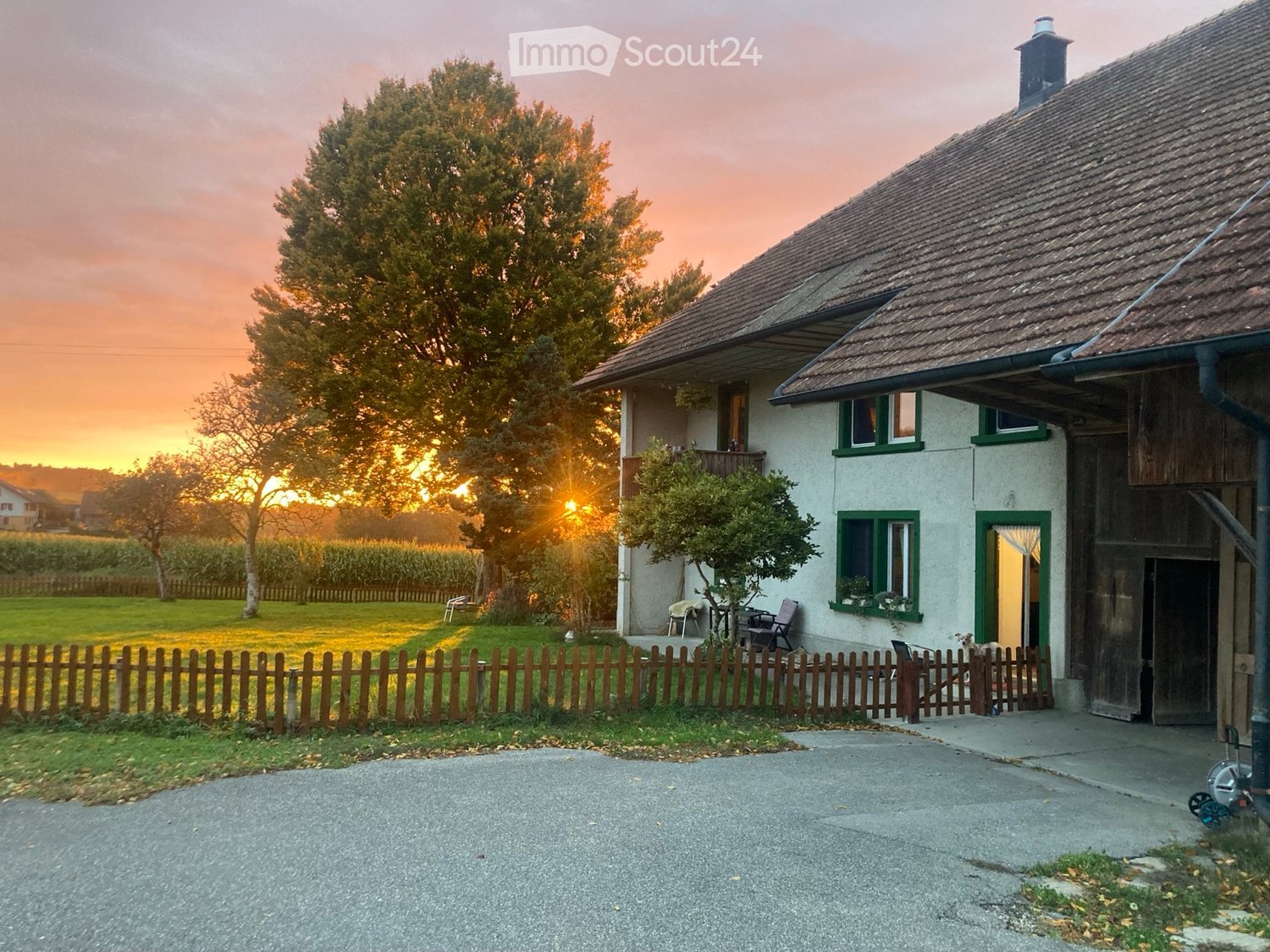 Two-story house with white walls, green-framed windows, and a wooden garage. The house is surrounded by a wooden fence, a grassy lawn, and trees, with a sunset view in the background.