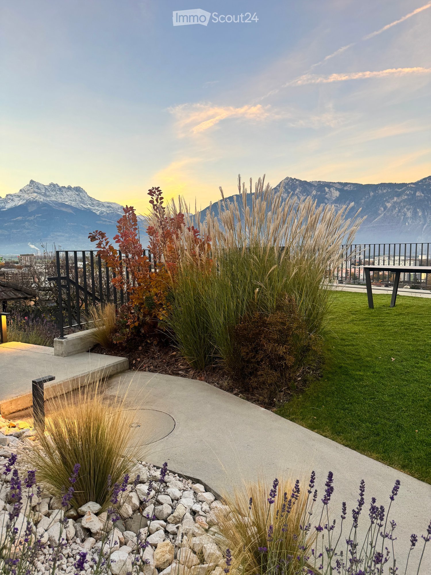 Well-maintained garden with tall grasses, shrubs, lavender plants, and a concrete pathway leading to a grassy area with a table and railing overlooking a mountain view.