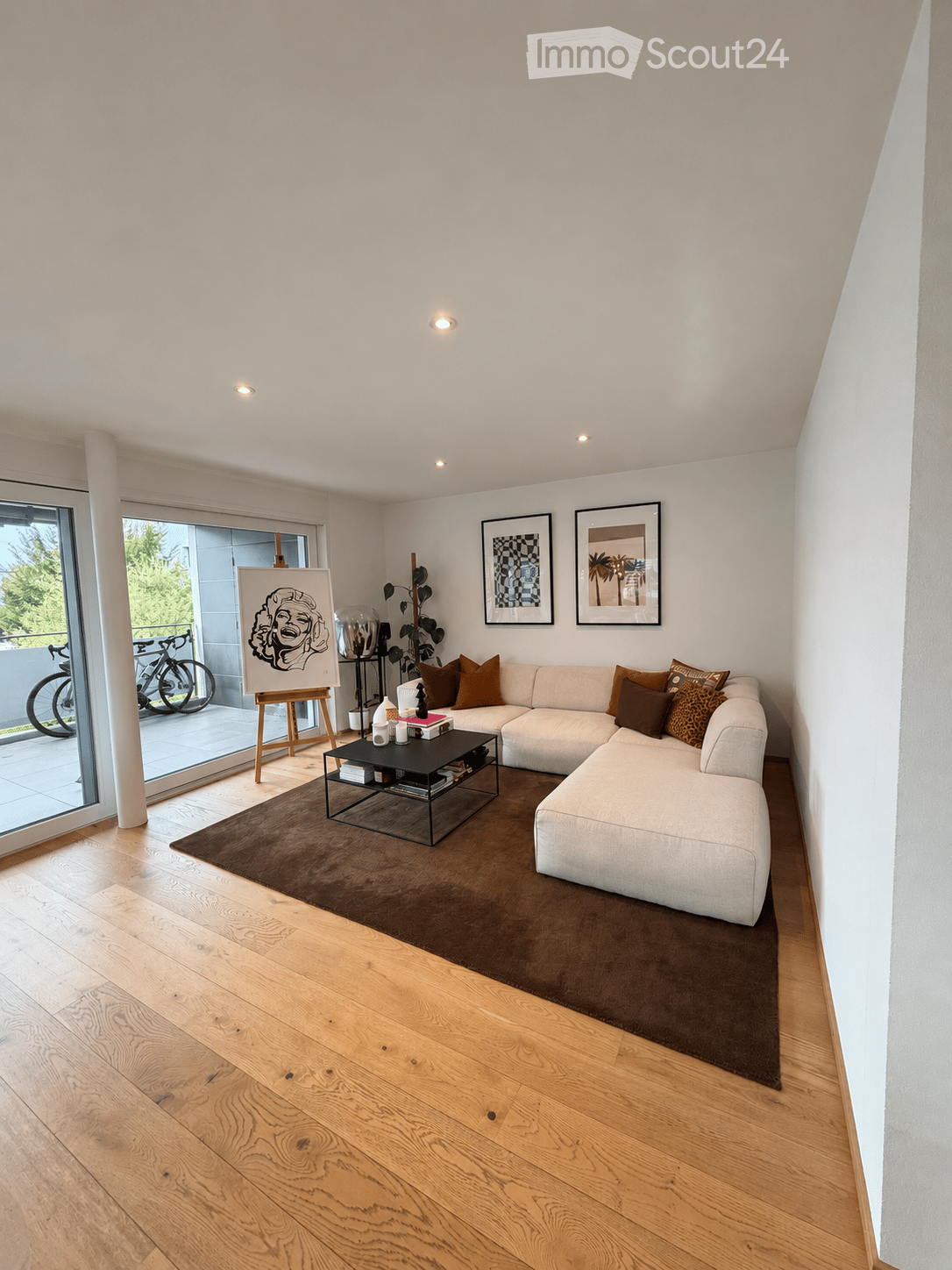 Modern living room with light wooden flooring, a large sectional sofa, a coffee table, and artwork on the walls. The room features large glass doors leading to a balcony with bicycles outside.