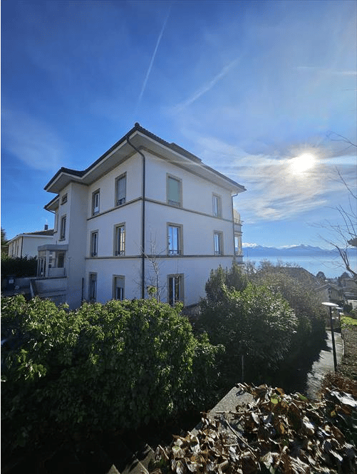 A multi-story residential building with white walls, multiple windows, and a view of mountains and a body of water in the background.