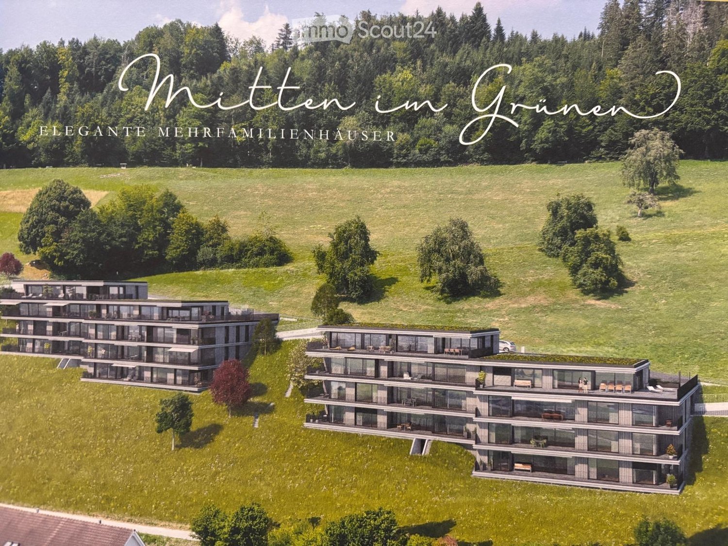 Aerial view of three modern multi-family houses with large windows and balconies, surrounded by green fields and trees.