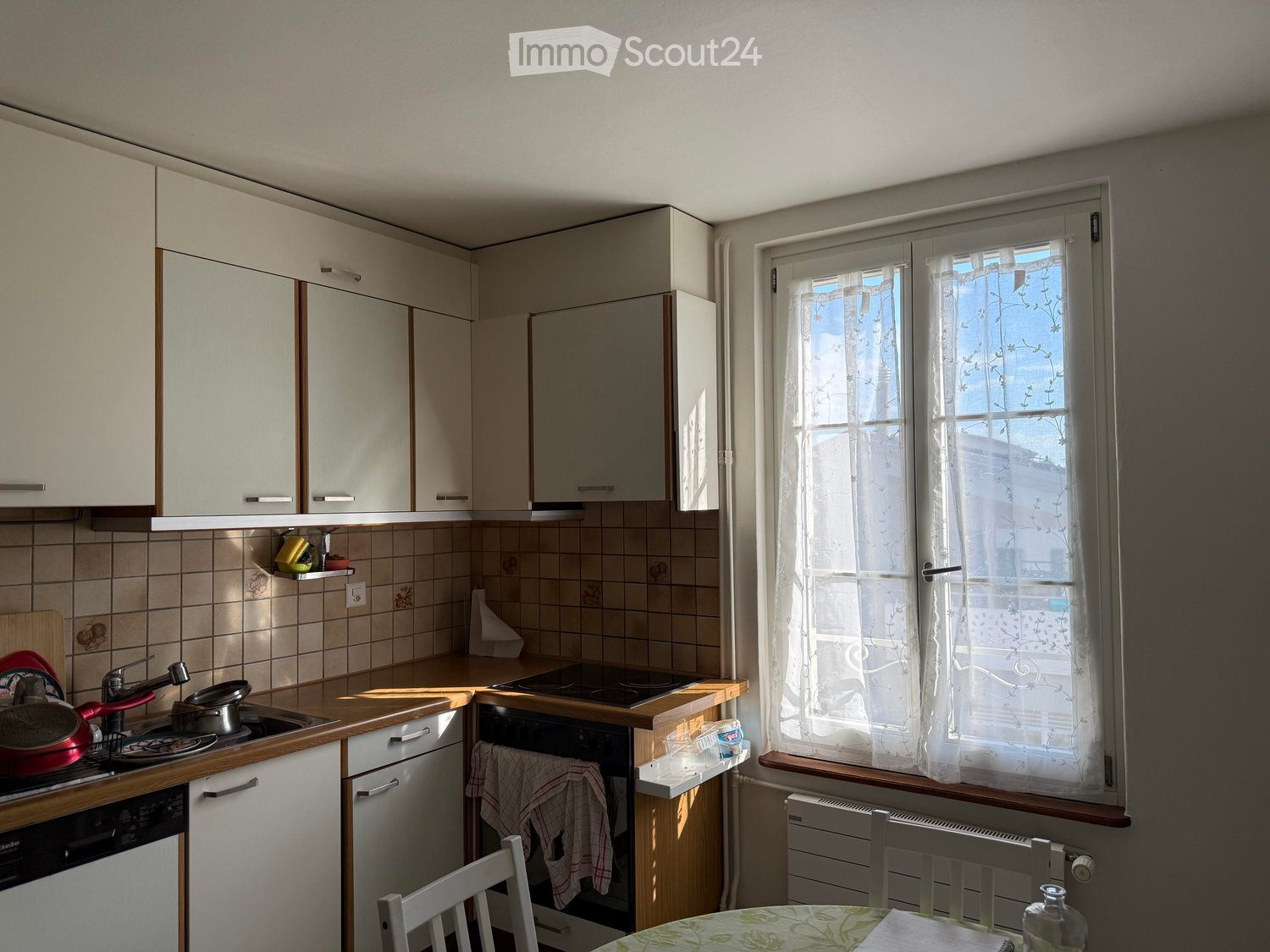 A small kitchen with white cabinets, a tiled backsplash, and a window with sheer curtains. The counter has a sink, stove, and various kitchen items, and there is a small dining area with a table and chairs.