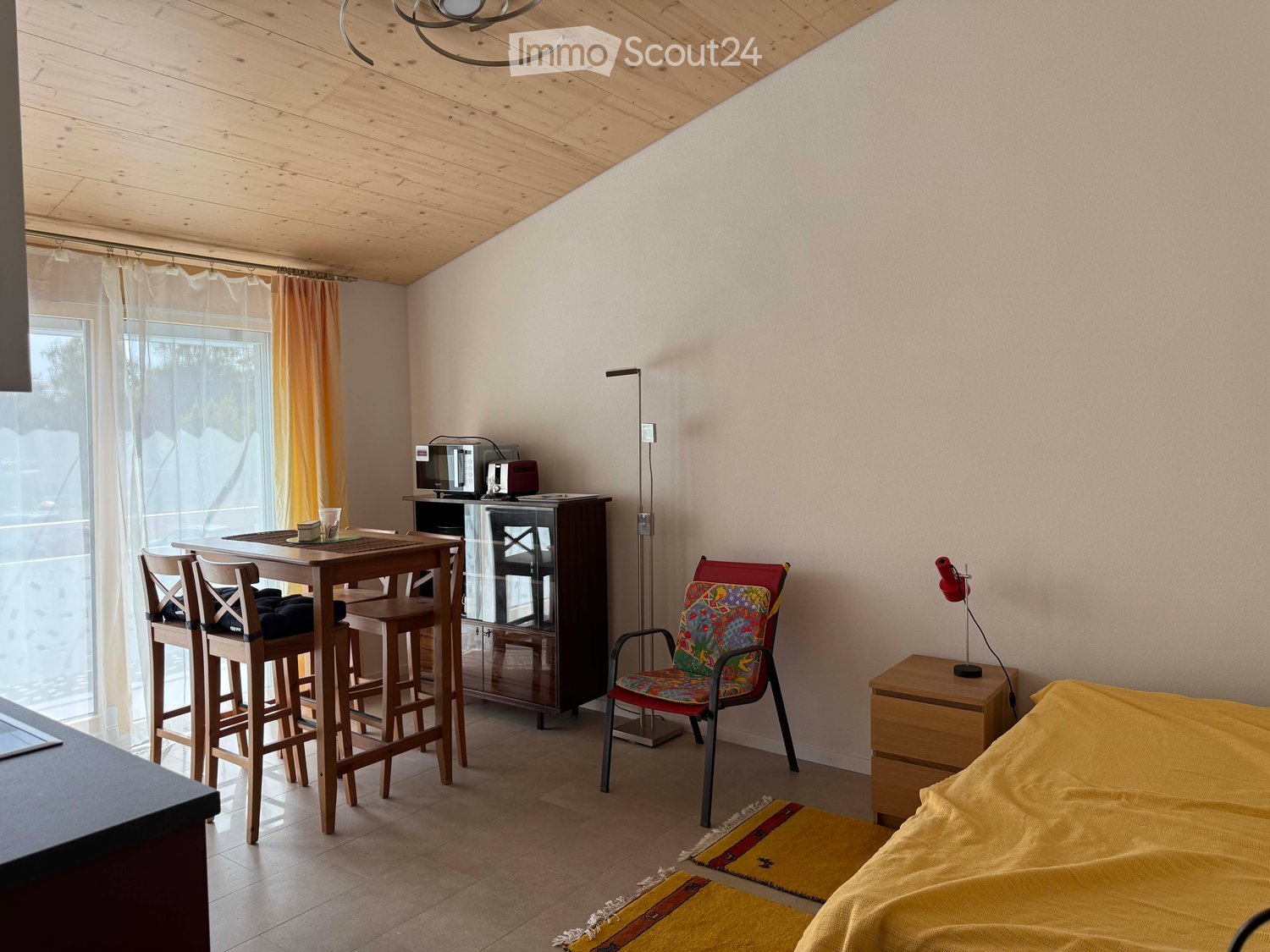 A cozy, well-lit room with a bed, a small dining area, a colorful chair, and a modern lamp. The room features wooden furniture and a warm, inviting atmosphere.