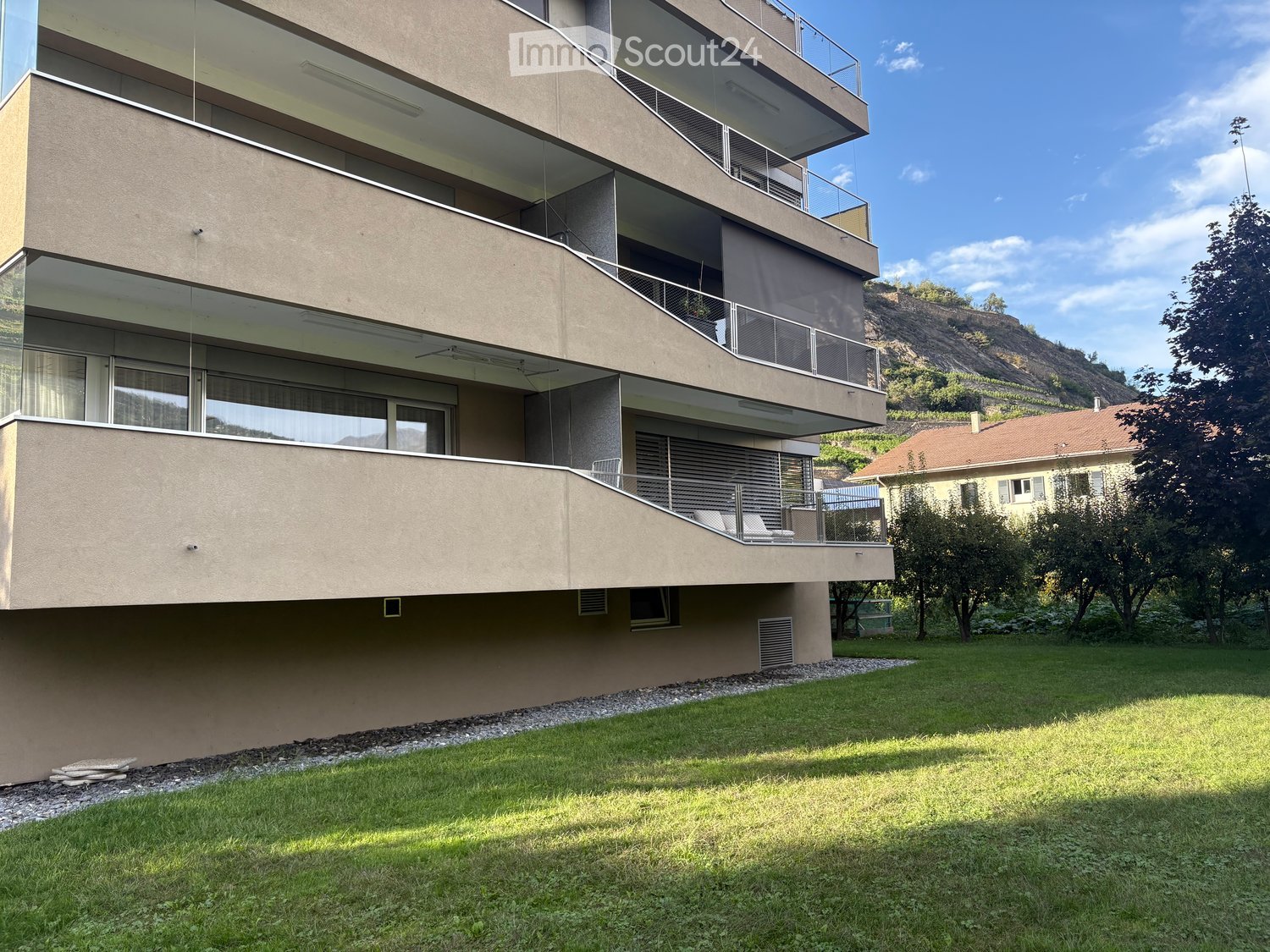 Modern multi-story building with large windows and balconies, surrounded by a well-maintained lawn and trees.