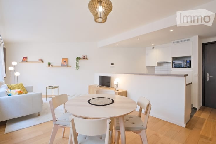 Modern, open-plan living and dining area with a white sofa, small round dining table with four chairs, a television on a stand, and a compact kitchen area with white cabinets and a microwave. Shelves with decorative items and a pendant light are also visi