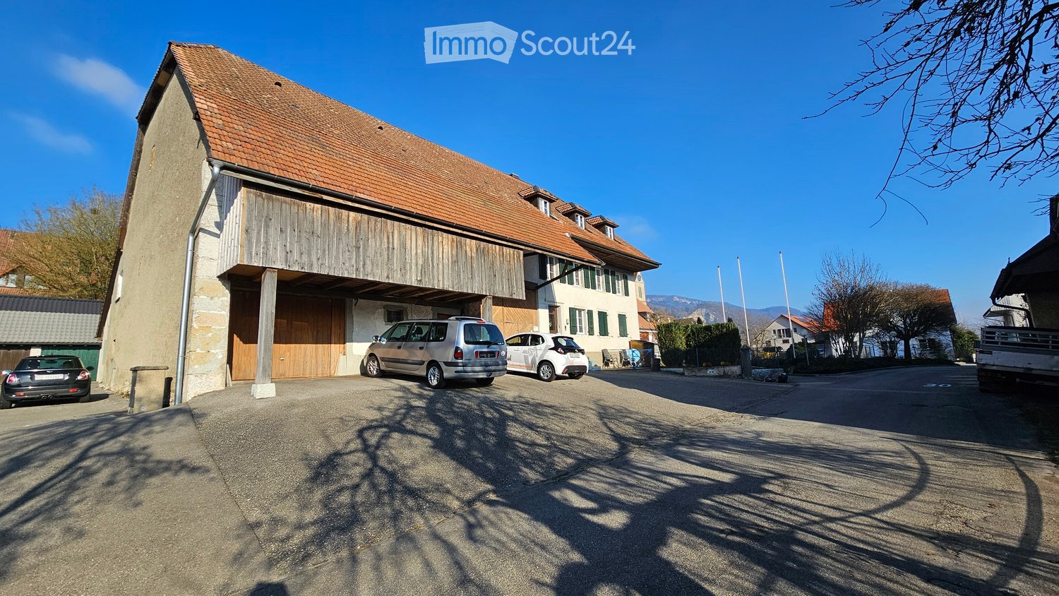 Two-story house with a red-tiled roof, stone and wooden exterior, and a covered driveway with several parked cars.