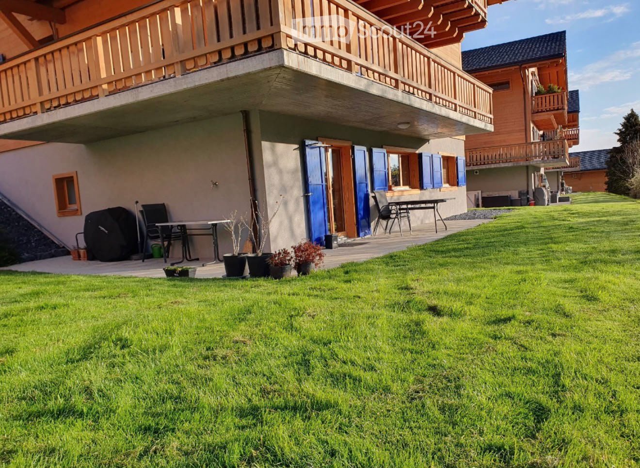 Lush green lawn in front of a two-story wooden house with balconies, outdoor furniture, and potted plants.