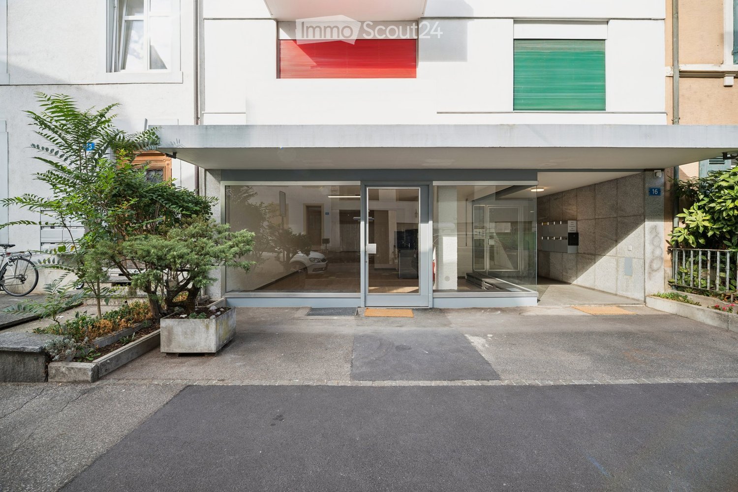 Modern multi-story building with a raised ground floor entrance, large glass doors, and a small landscaped area with trees and shrubs.