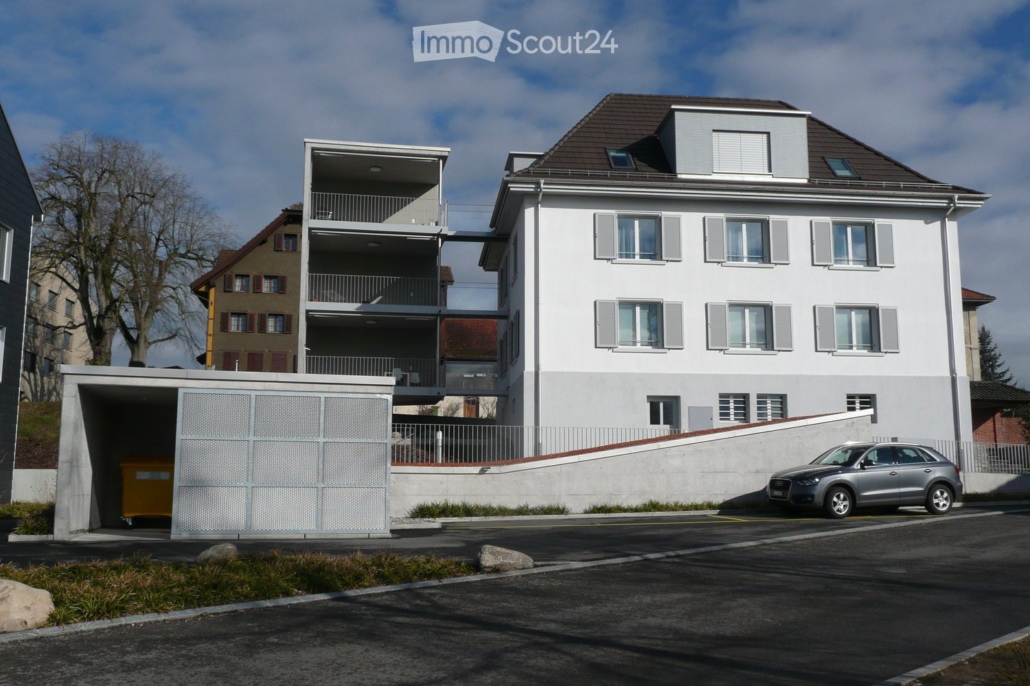 Modern two-story residential building with white facade, grey shutters, and a balcony on the upper level. A covered garage entrance is visible on the left, and a silver SUV is parked in front of the building.