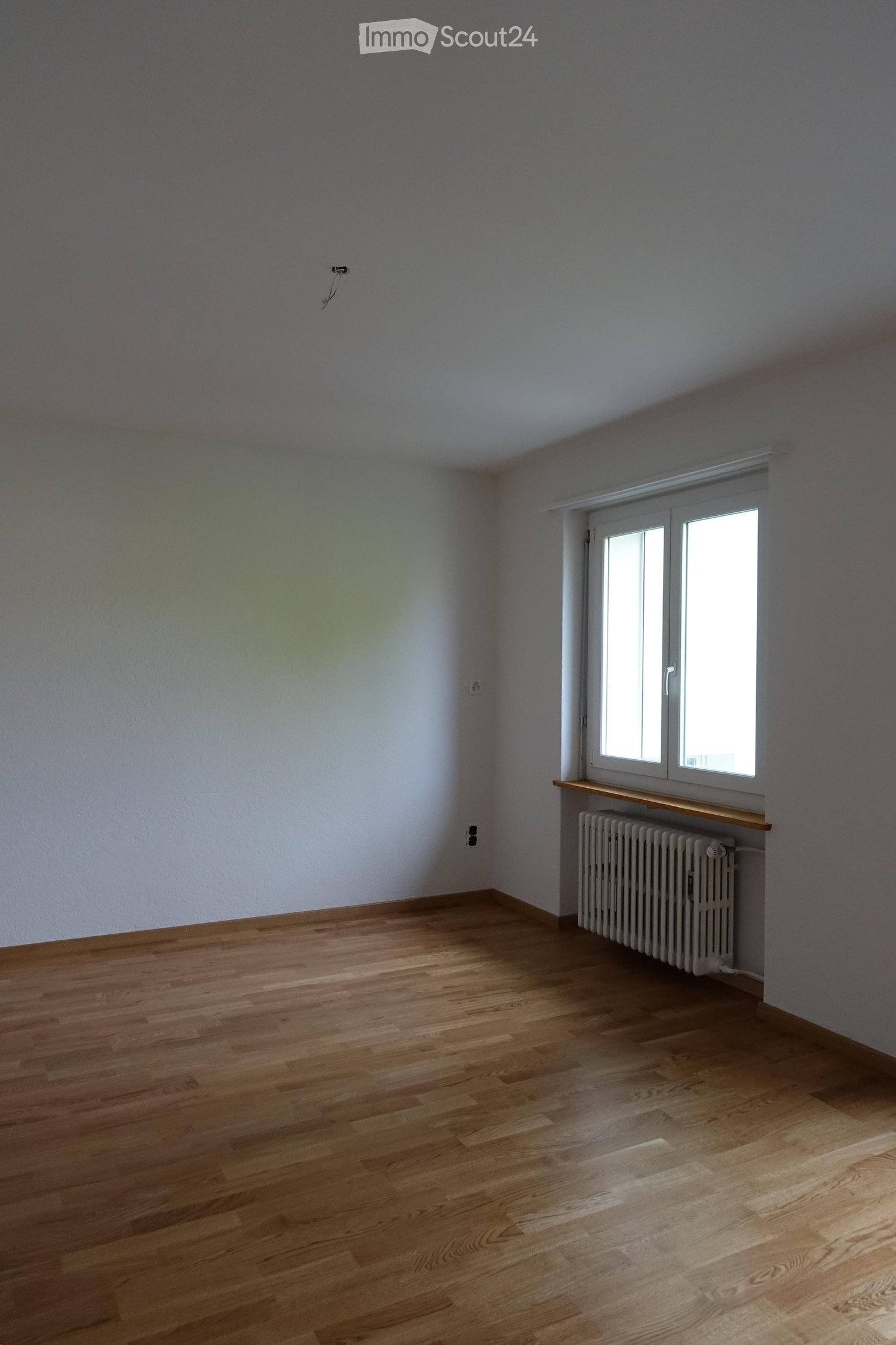 Empty room with wooden flooring, white walls, and a window with a radiator below it.
