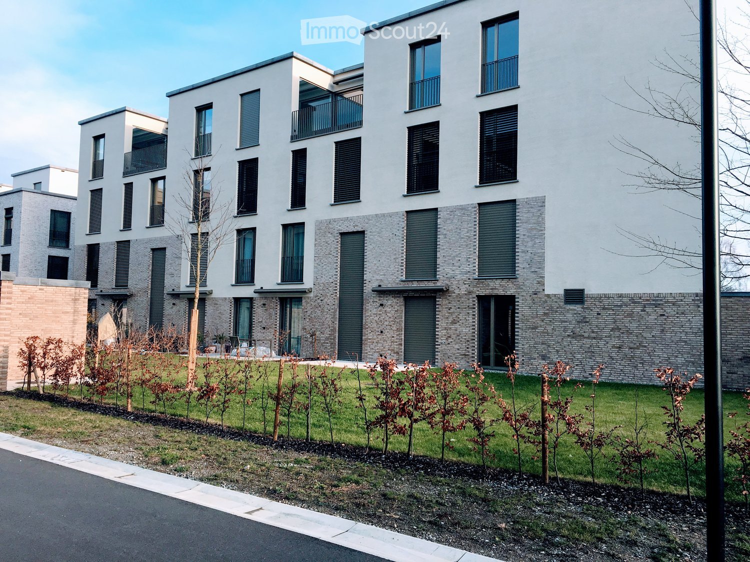 Modern multi-story residential building with white facade, large windows, and balconies. The building features a mix of brick and concrete elements, surrounded by a landscaped garden area with small trees and shrubs.