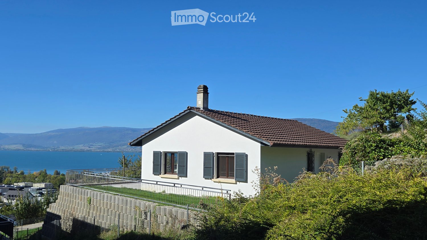 Single-story house with white walls, brown tiled roof, and green shutters. Located on a raised ground floor with a stone retaining wall and metal railing, overlooking a body of water and distant mountains.