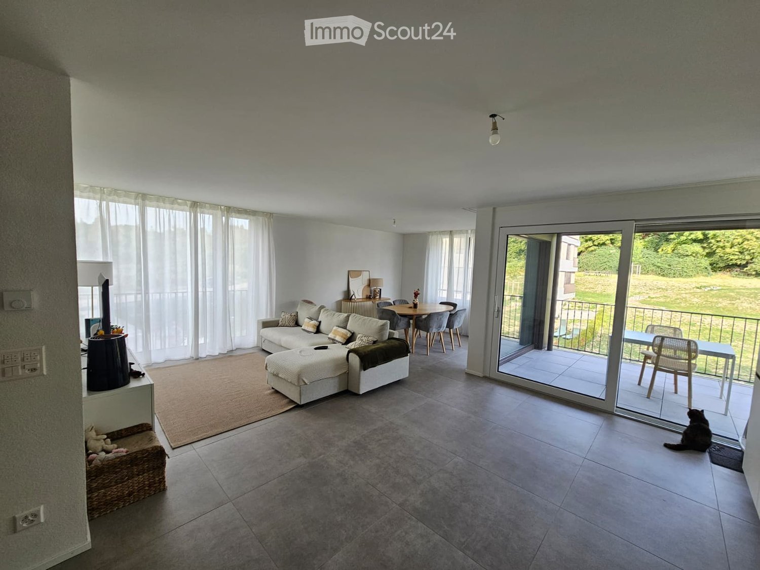 Modern living room with a grey sectional sofa, ottoman, coffee table, dining area with table and chairs, large windows with white curtains, and a cat sitting on the balcony.