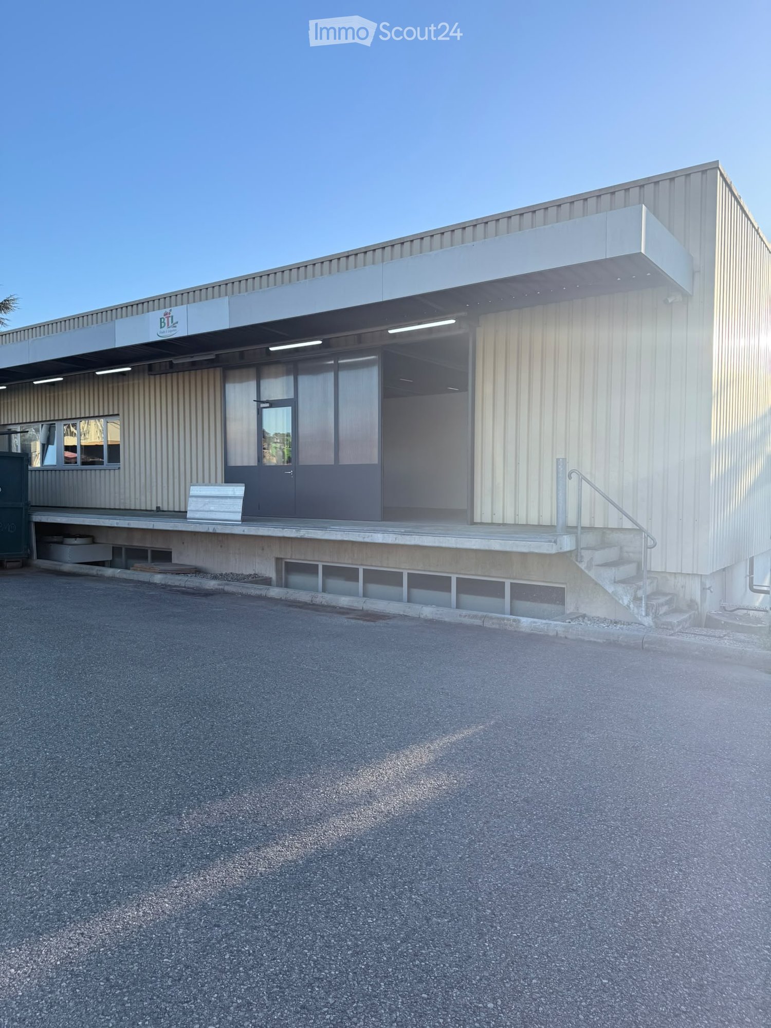 Modern industrial-style building with a metal facade, large windows, and a covered entrance area. Features a concrete staircase leading to the entrance and a small bench near the entrance.