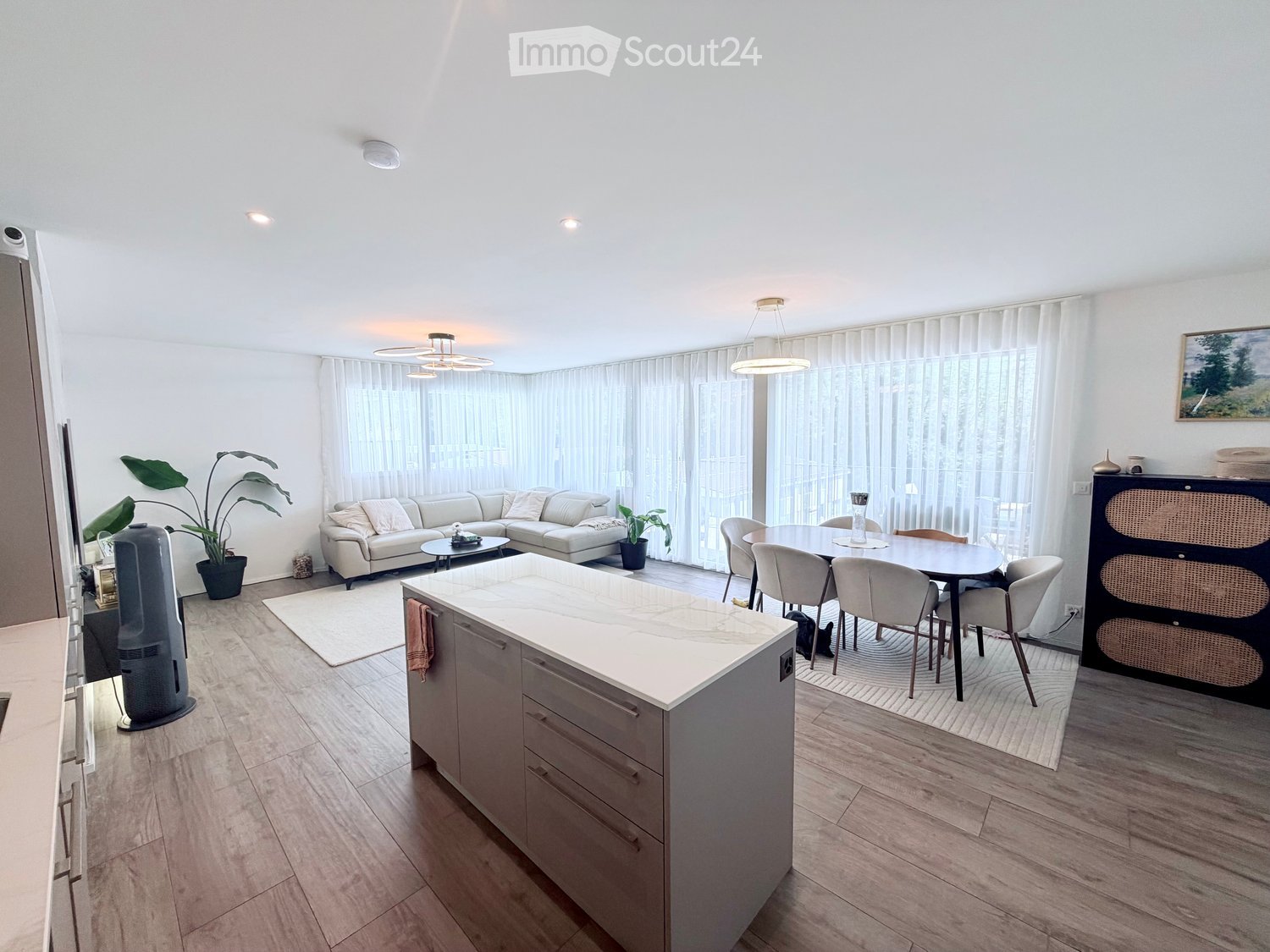 Open-plan living and dining area with a white sofa, coffee table, dining table with chairs, kitchen island, and large windows with curtains.