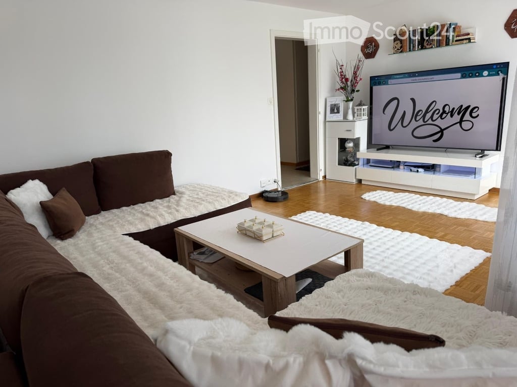 Modern living room with a brown sectional sofa, white shaggy rug, white coffee table, flat-screen TV displaying "Welcome", and decorative shelf with books and a vase.
