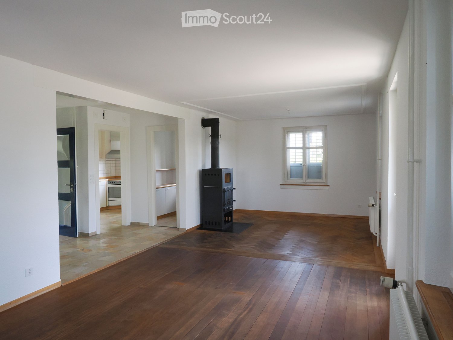 Spacious room with wooden flooring, white walls, and a wood stove in the center. A radiator is visible on the right side, and a doorway leads to a kitchen area with white cabinets and appliances.