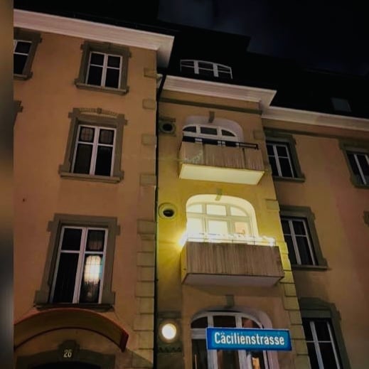 Multi-story residential building with illuminated balconies and windows, featuring a street sign reading "Cäcilienstrasse".