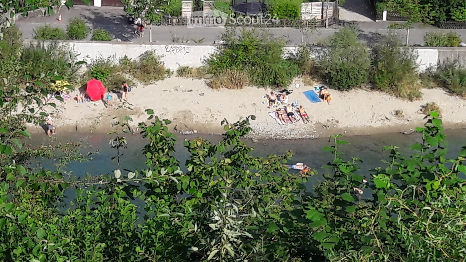 A sandy beach area with people relaxing, sunbathing, and swimming in the water. The area is surrounded by greenery and a concrete wall with graffiti.