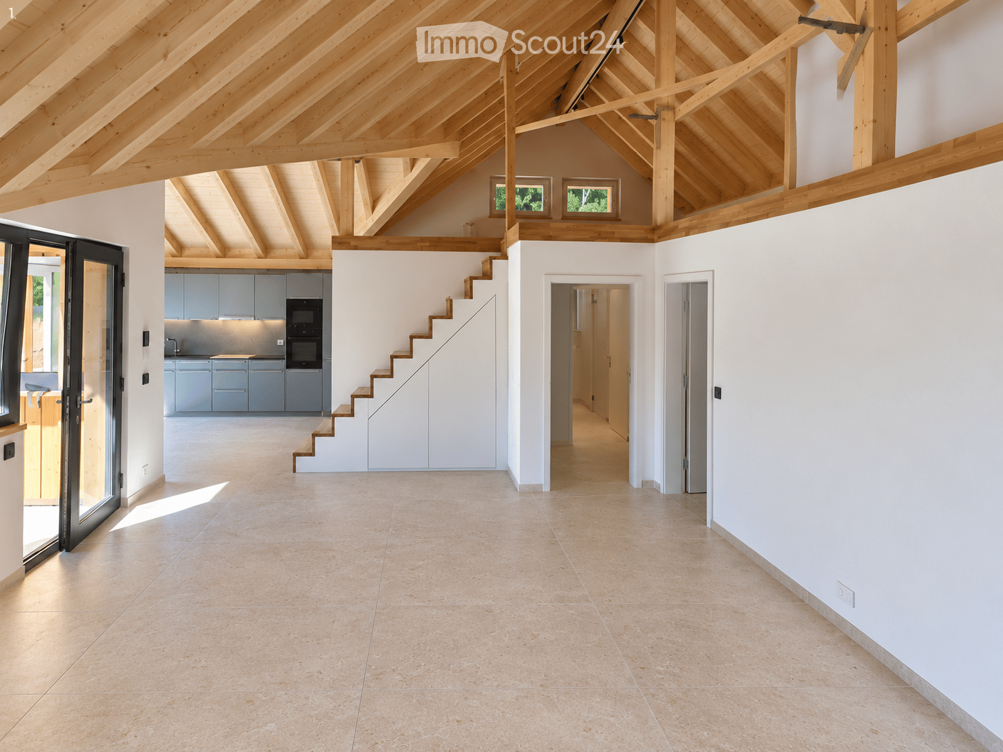 Open-plan space with exposed wooden beams, tiled floor, and white walls. Features a staircase leading to an upper level, a kitchen area with cabinets and appliances, and large windows allowing natural light.