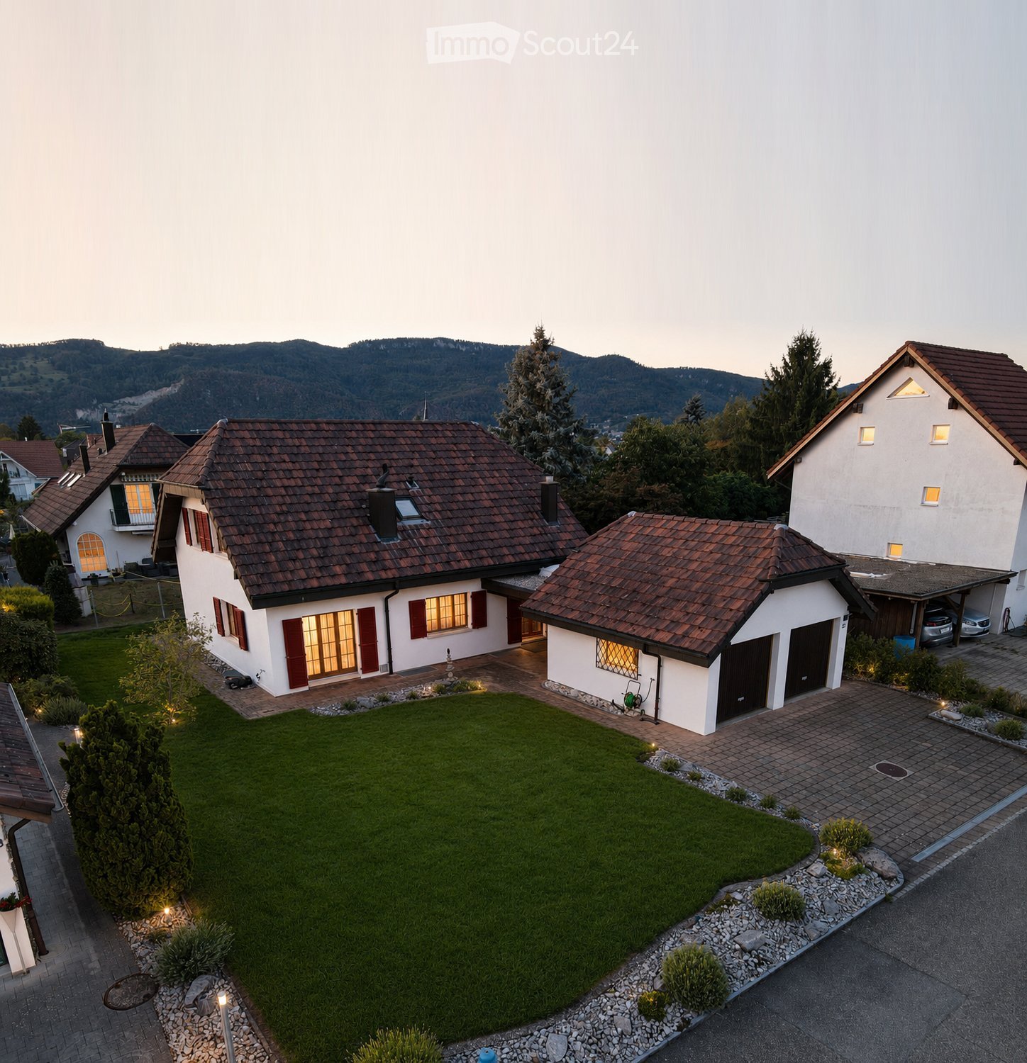 A two-story house with a red-tiled roof, white walls, and multiple windows, surrounded by a well-maintained lawn and garden, with a driveway and garage visible.