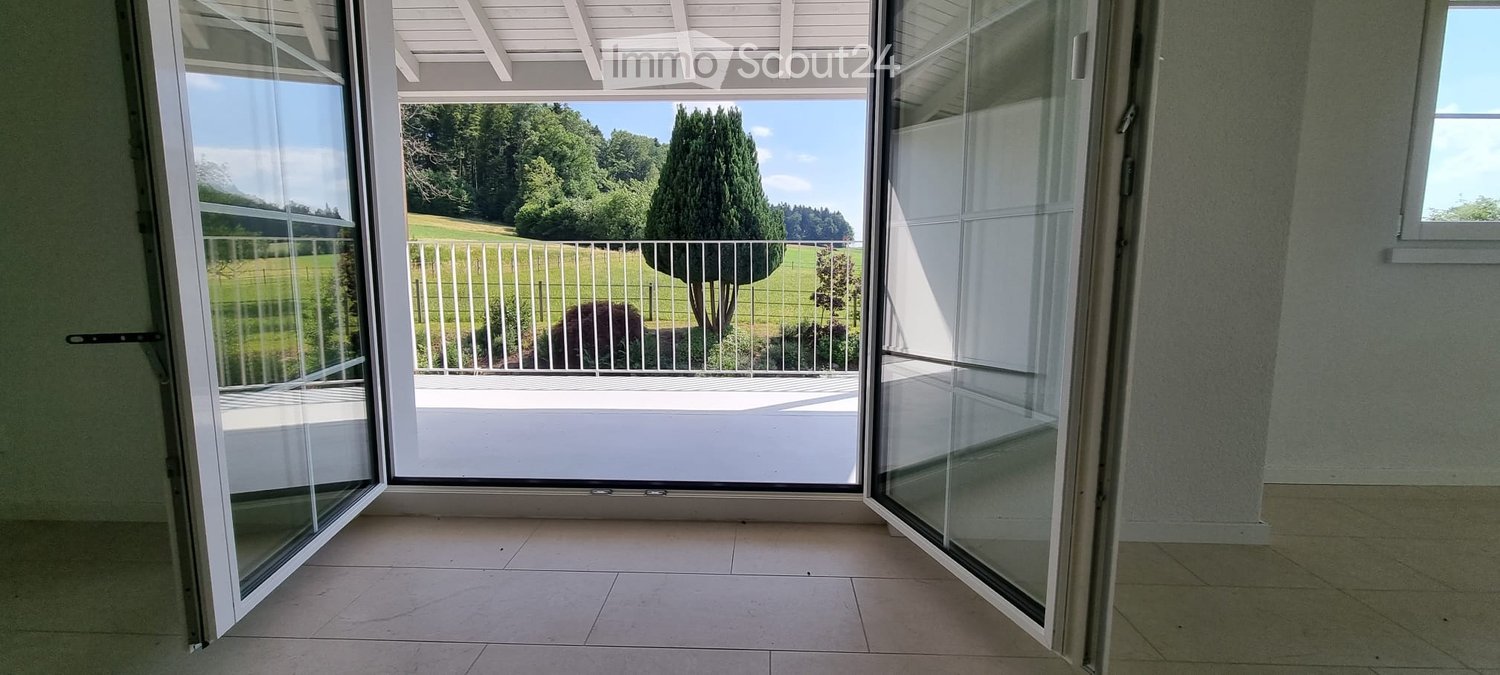Open sliding glass doors lead to a balcony with a view of a green field and trees.