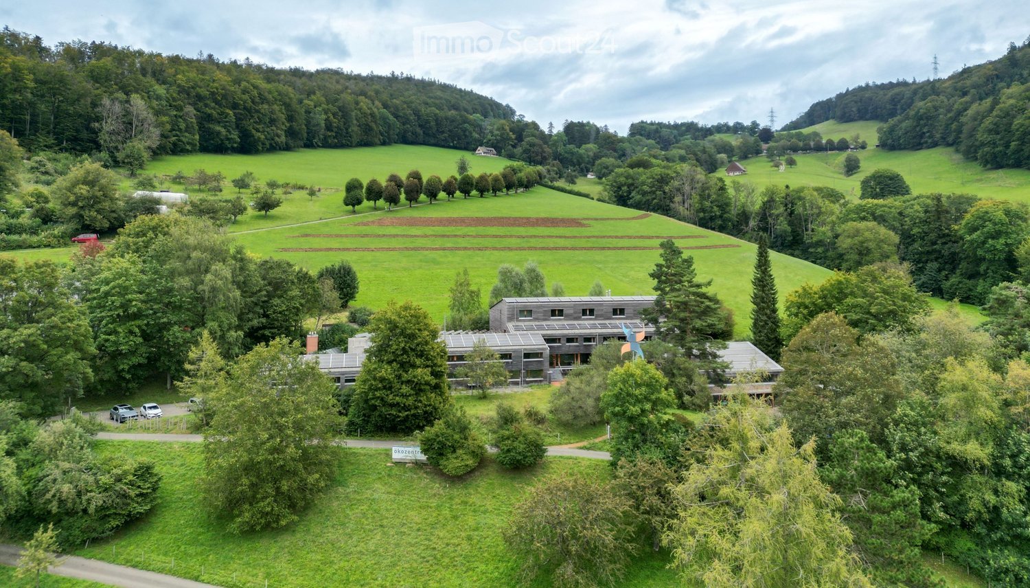 A modern, low-rise building surrounded by lush green fields and trees, with a parking area and a few scattered vehicles.