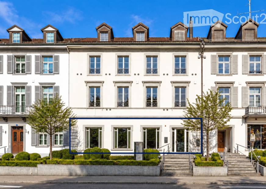 A large, elegant white building with multiple stories, numerous windows, and a classic architectural style. The building features symmetrical design, decorative shutters, and a well-maintained garden area in front.
