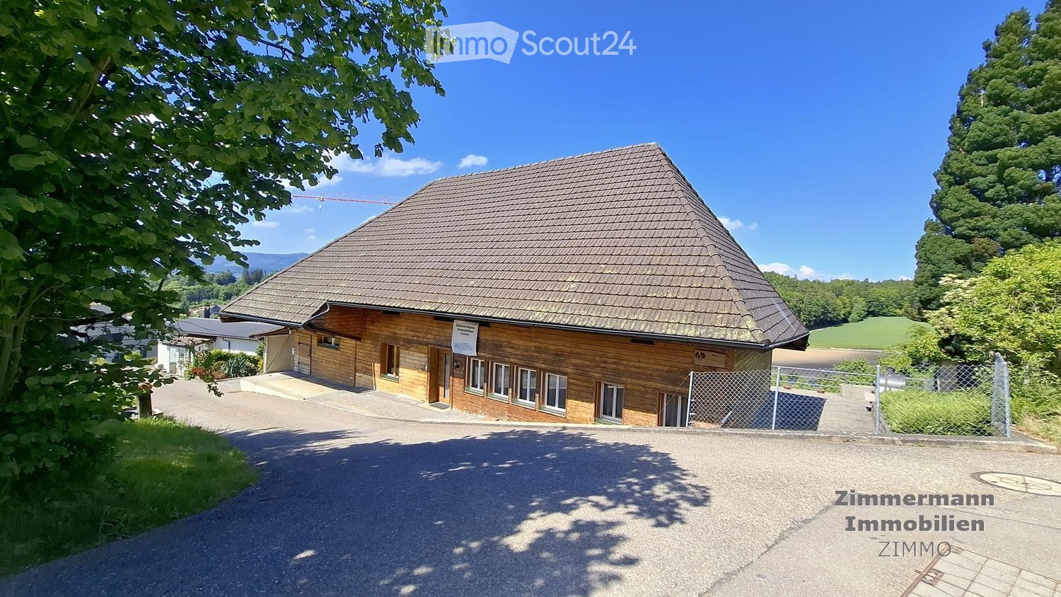 Single-story wooden house with a pitched roof, surrounded by greenery and a gravel driveway.