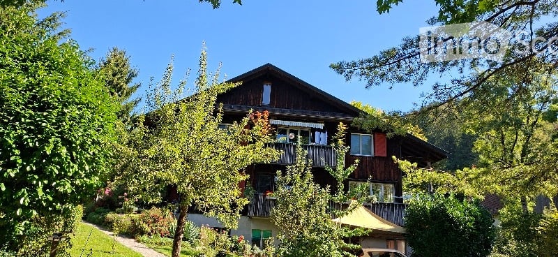 Two-story wooden house with balconies, surrounded by lush greenery and trees.