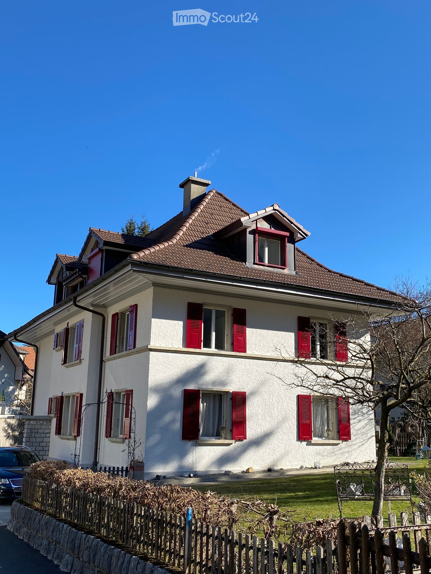 Two-story house with white exterior, red shutters, and a tiled roof. Features a chimney, multiple windows, and a small garden with a bench and tree.