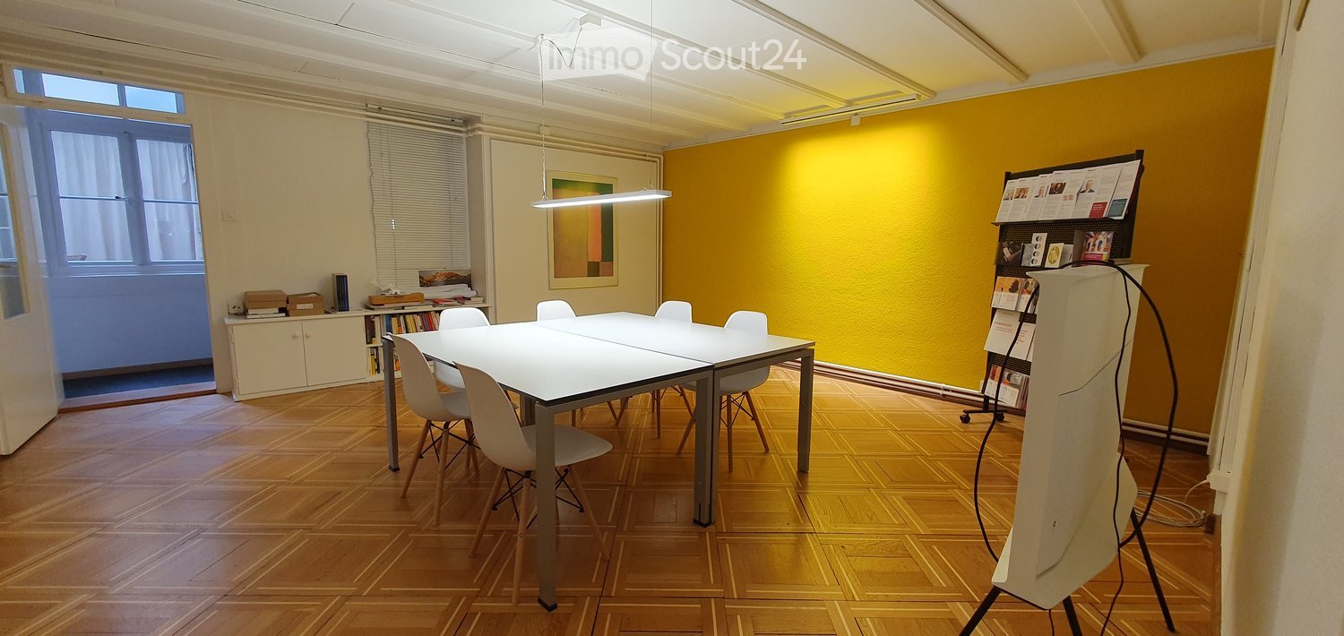 Bright room with wooden flooring, white table and chairs, a cabinet with books, and a screen on a stand.