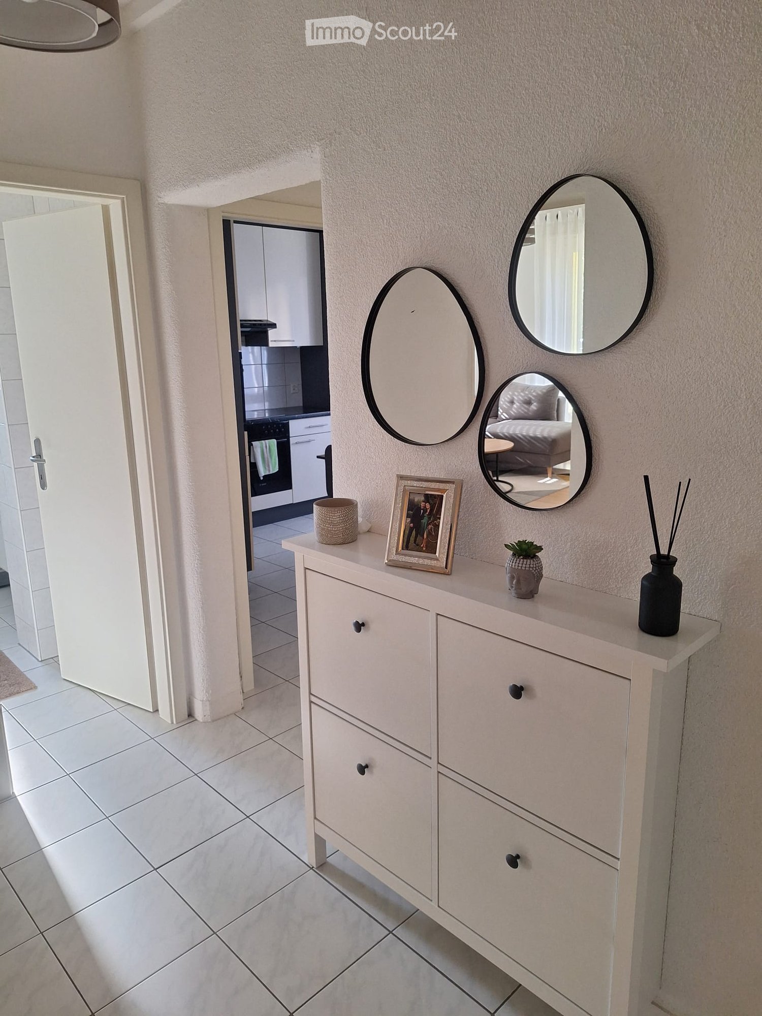 Hallway with white tiled floor, light-colored walls, and a small console table with decorative items, mirrors, and a view into a kitchen and bedroom area.