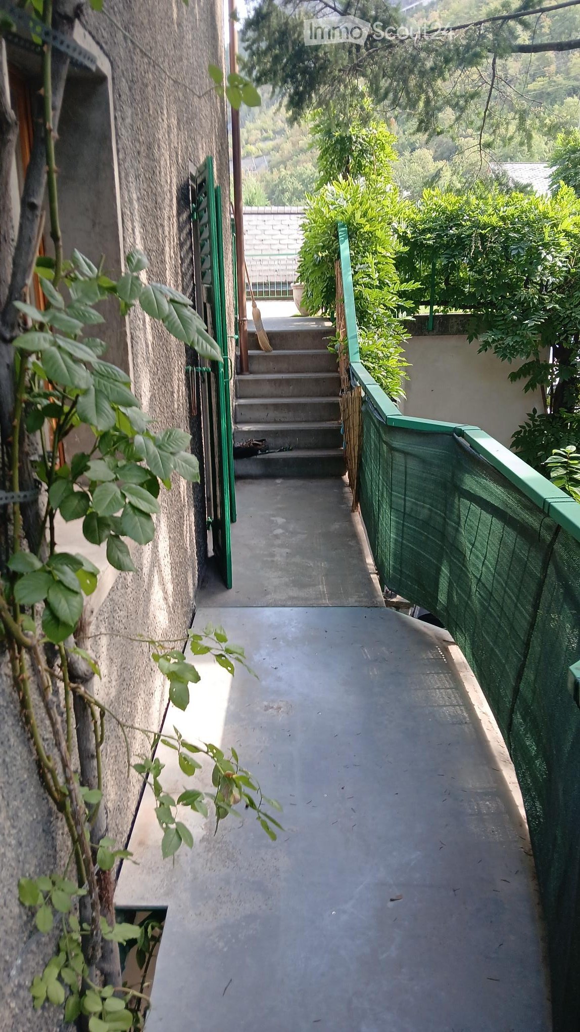 A narrow concrete pathway bordered by green railings, leading to a set of stairs and a small outdoor area with plants and a view of greenery.