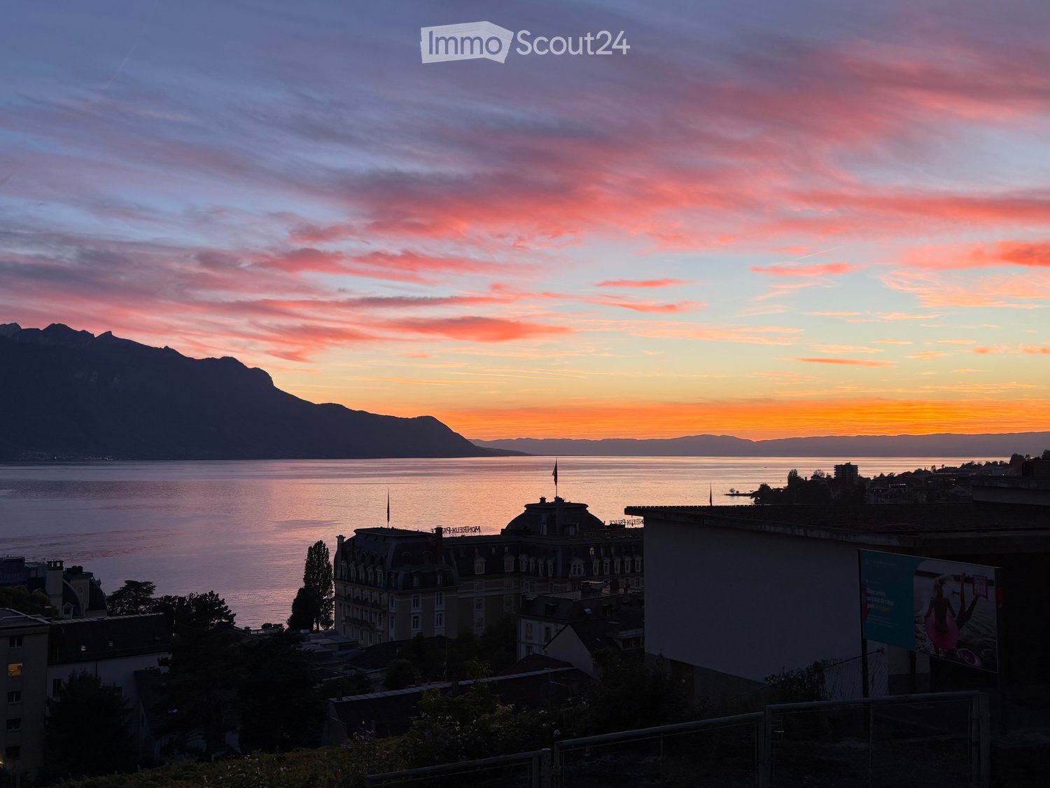 View from a balcony or terrace overlooking a lake and mountains at sunset, with city buildings in the foreground.