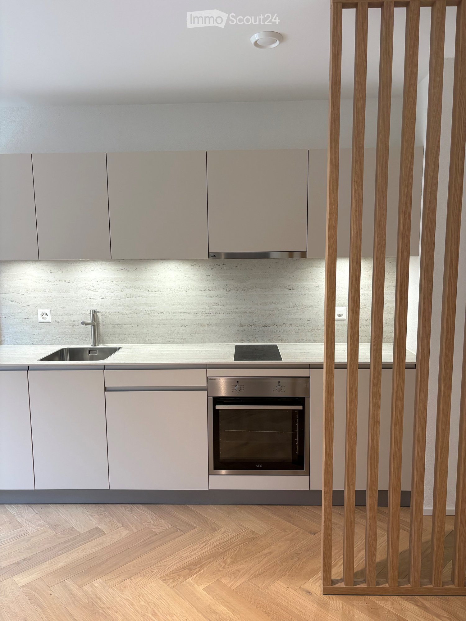 Modern kitchen with light grey cabinets, a stainless steel sink, a built-in oven, and a sleek countertop. The floor is made of light wooden planks arranged in a herringbone pattern.