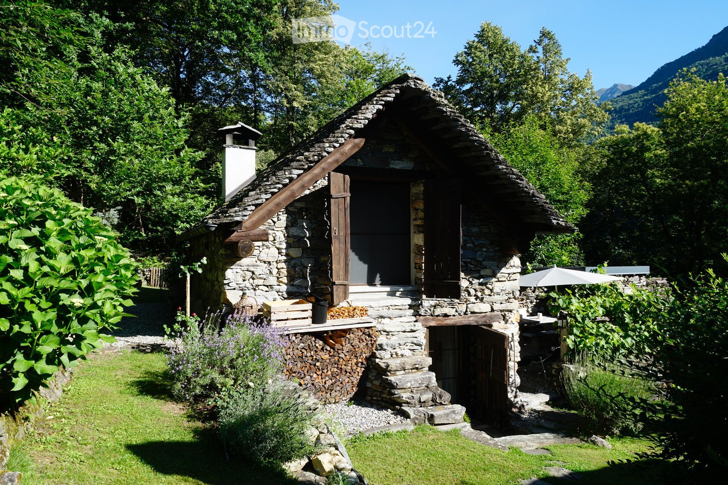 A rustic stone cottage with a steep, slate roof, surrounded by lush greenery and a scenic mountain backdrop. Features include a chimney, wooden shutters, a small porch, and a neatly stacked woodpile.