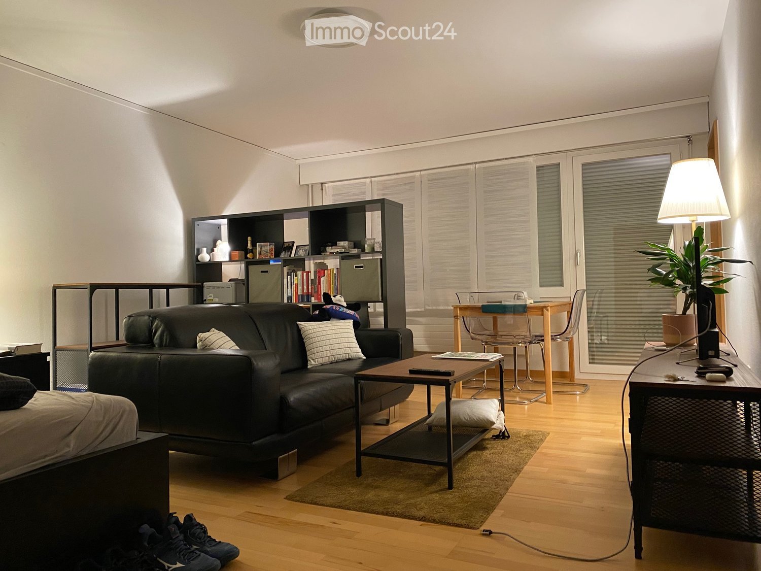 Modern living room with a black leather sofa, coffee table, bookshelf, small table with chairs, and a potted plant near the window.