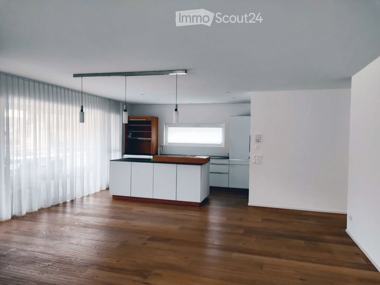 Modern kitchen with white cabinets, wooden countertop, and pendant lights. Features a wooden shelf, a sink with a faucet, and a refrigerator. The space is illuminated by natural light from a window covered with sheer curtains.