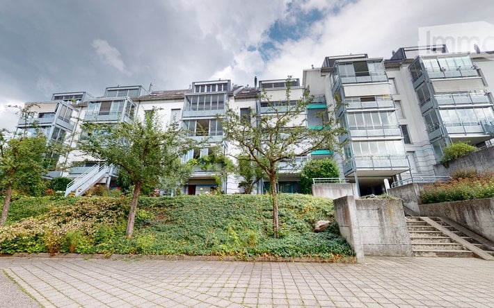 Modern multi-story residential building with large windows and balconies, surrounded by greenery and a paved walkway.